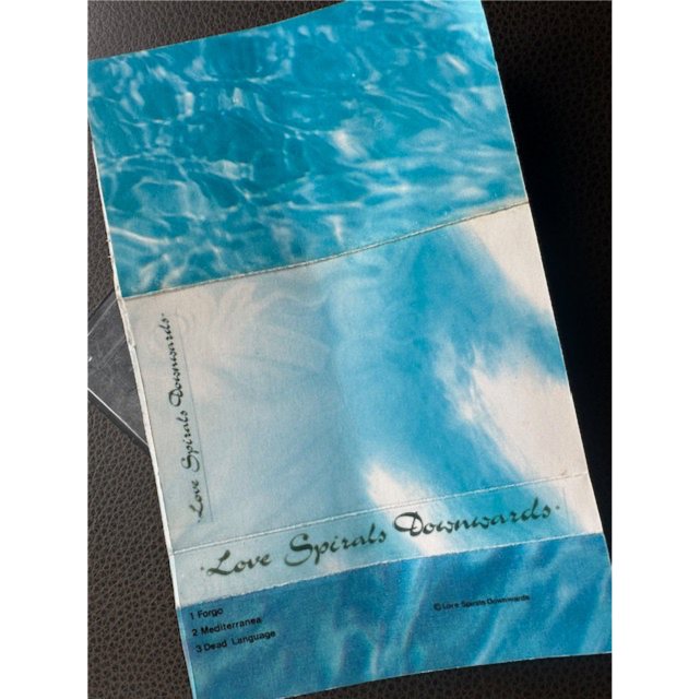 The blue water-pattern cover of the original Love Spirals Downwards demo cassette, hand-lettered with the band name and the songs Forgo, Mediterranea and Dead Language
